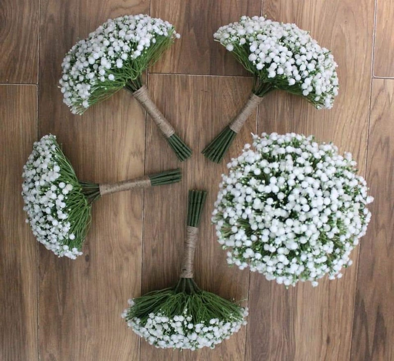 May include: Five white baby's breath bouquets tied with twine, arranged in a circle on a wood floor.