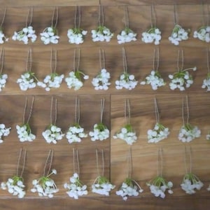 May include: A set of 48 white flower hair pins arranged in rows on a wooden surface. The pins are made of white flowers with green stems and are attached to a thin wire.