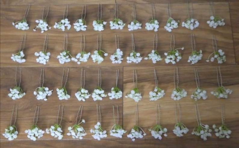 May include: A set of 48 white flower hair pins arranged in rows on a wooden surface. The pins are made of white flowers with green stems and are attached to a thin wire.