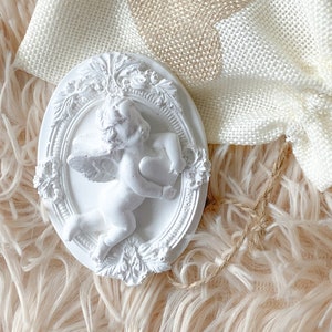 Peut inclure: Figurines d'ange en céramique blanche avec des ailes, tenant un cœur, dans un cadre ovale aux détails ornés. La figurine est sur un sac en toile de jute blanche avec un motif en forme de cœur.