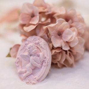 Op de afbeelding: Een roze keramiek cherubijn figuur dat een hart vasthoudt, ingelijst in een ovaal met sierlijke details, omringd door roze hortensia bloemen.