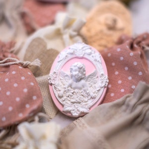 Può includere: Un ornamento ovale in ceramica rosa con un designo di cherubino bianco. Il cherubino tiene un fiore e ha le ali. L'ornamento è circondato da piccole borse di stoffa.
