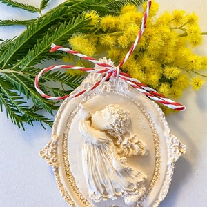 May include: A white ceramic ornament shaped like an oval frame with a 3D sculpted image of a girl in a dress holding a flower. The ornament is decorated with intricate floral details and is tied with a red and white string.