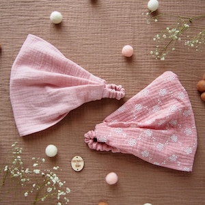 Op de afbeelding: Twee roze haarbanden met elastiek, één effen en één met een wit bloemmotief. De haarbanden zijn gemaakt van een zachte, getextureerde stof. De achtergrond bevat decoratieve elementen zoals kleine ballen en bloemen.