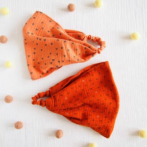 May include: Two fabric headbands with elastic back bands. One headband is light orange with a small black heart pattern, and the other is a darker burnt orange with a small black star pattern. Both feature a gathered, textured fabric design.