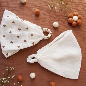 May include: Two white fabric headbands with elastic scrunchie-style closures. One headband features a repeating pattern of small brown hearts, while the other is plain white. Both are made of a soft, crinkled muslin-like textile material.