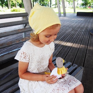 Peut inclure: Une jeune personne portant un foulard jaune et une robe en dentelle blanche, tenant un jouet en forme de canard en peluche. Le canard est blanc avec une tête grise et des détails jaunes. La personne est assise sur un banc gris à l'extérieur.