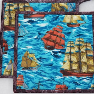 May include: Two square pot holders with a nautical theme. The fabric features a pattern of sailing ships on a blue ocean. The ships are in shades of red, yellow, and brown. The edges are trimmed with a dark brown fabric.