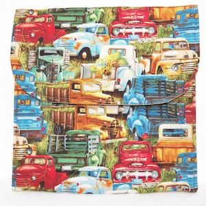 May include: A fabric item with a flap closure, featuring a repeating pattern of vintage pickup trucks in various colors like red, blue, and yellow. The trucks are set against a backdrop of green grass and a light background.