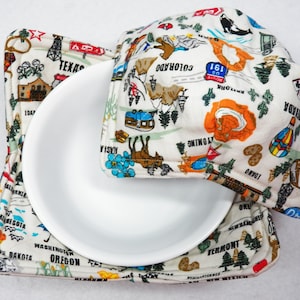 May include: A white bowl with a white and colorful fabric pot holder with a map of the United States printed on it. The pot holder has the names of states printed on it, including Texas, Colorado, Wyoming, Idaho, Minnesota, South Dakota, Oregon, Washington, Vermont, and New Mexico.