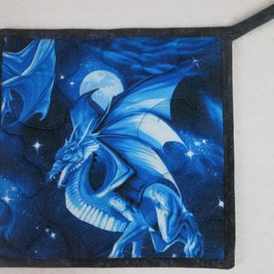 May include: Square blue and black quilted potholder featuring a detailed illustration of a blue dragon with outstretched wings against a starry night sky with a full moon. The potholder has a dark gray border and a hanging loop.