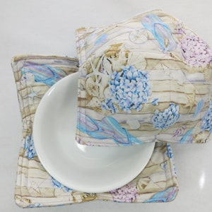May include: A set of two beige and blue fabric pot holders with a beach-themed design featuring seashells, starfish, and hydrangeas. The pot holders are quilted and have a square shape.