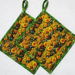 May include: Two square quilted pot holders featuring a vibrant floral pattern with yellow and orange flowers on a dark background. Each pot holder is finished with a green fabric border and includes a small fabric loop at the top corner for hanging.