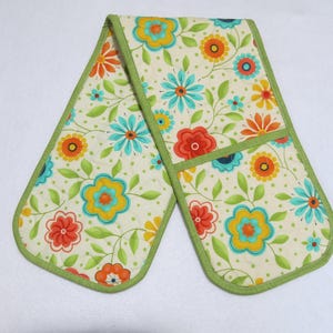 May include: A double oven mitt featuring a cream-colored background with a repeating pattern of colorful flowers in shades of orange, yellow, and teal, accented by green leaves and small dots, all finished with a solid green fabric trim border.
