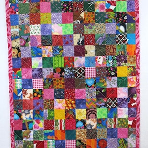 May include: A rectangular patchwork quilt composed of numerous small, multi-colored fabric squares featuring diverse patterns, including florals, geometric shapes, and sports motifs, all bordered by a pink fabric trim with a swirling design.
