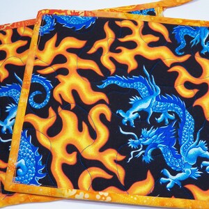May include: Two orange and yellow quilted pot holders with a black background and blue dragon designs. The dragons are surrounded by flames.