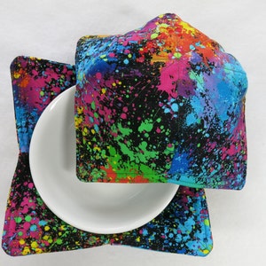 May include: A set of two colorful bowl cozies with a black background and a splatter paint design. The cozies are made of fabric and are quilted.