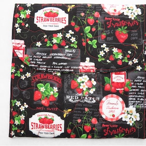 May include: Black fabric with a repeating pattern of red strawberries, white flowers, and green leaves. The fabric has a quilted texture and features several labels with text that reads "Sweet & Juicy Strawberries Pick Your Own", "Homemade Strawberry Jam", and "Red Patch Strawberry Jam".
