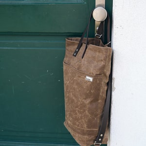 Pu&ograve; includere: Una borsa tote in tela marrone con cinturini in pelle e una piccola toppa in pelle marrone con la scritta "Riddel". La borsa &egrave; appesa a una maniglia della porta.