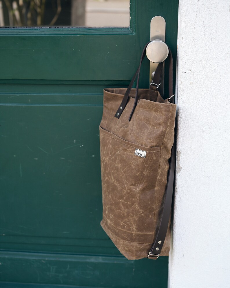 Pu&ograve; includere: Una borsa tote in tela marrone con cinturini in pelle e una piccola toppa in pelle marrone con la scritta "Riddel". La borsa &egrave; appesa a una maniglia della porta.