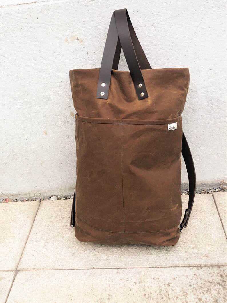 Pu&ograve; includere: Una borsa tote in tela marrone con manici in pelle e una tracolla in pelle marrone. La borsa ha una grande tasca sul davanti e una piccola etichetta con la parola "Kikki" su di essa.