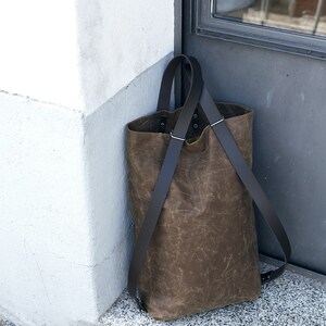 Pu&ograve; includere: Una borsa tote in pelle marrone con cinturini neri. La borsa &egrave; appoggiata su uno scalino di pietra accanto a un muro bianco.