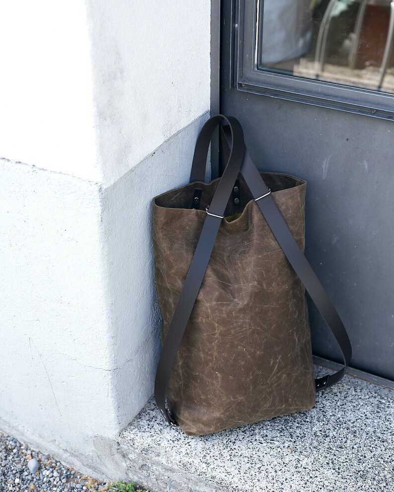 Pu&ograve; includere: Una borsa tote in pelle marrone con cinturini neri. La borsa &egrave; appoggiata su uno scalino di pietra accanto a un muro bianco.