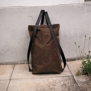 Pu&ograve; includere: Una borsa tote in tela marrone con cinturini in pelle nera. La borsa &egrave; appoggiata su una superficie di cemento.