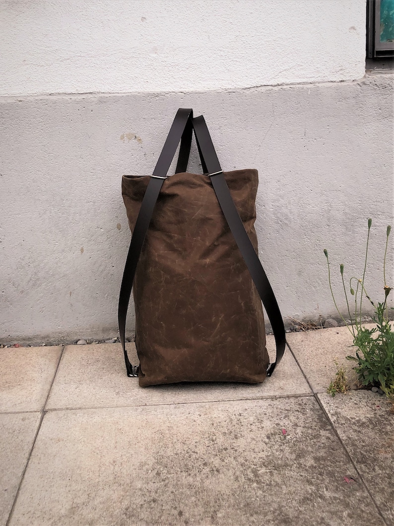 Pu&ograve; includere: Una borsa tote in tela marrone con cinturini in pelle nera. La borsa &egrave; appoggiata su una superficie di cemento.