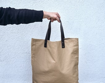 Può includere: Una borsa tote in tela beige con manici in pelle marrone e rivetti metallici. La borsa è tenuta da una mano contro un muro bianco.