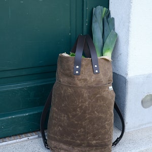 Pu&ograve; includere: Uno zaino in tela marrone con spalline in pelle e una grande tasca sul davanti. Lo zaino &egrave; pieno di verdure verdi.
