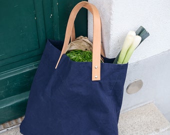 Può includere: Una borsa tote in tela blu navy con manici in pelle marrone e dettagli di cucitura bianchi. La borsa è piena di generi alimentari, tra cui verdure a foglia verde e cipolle bianche.