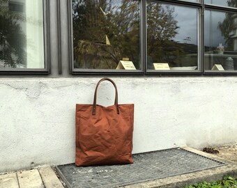Può includere: Una borsa tote in tela marrone con manici in pelle si trova su una superficie di cemento di fronte a un edificio con grandi finestre.