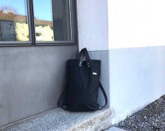 May include: A black canvas tote bag with leather straps and a small leather patch on the front. The bag is sitting on a step in front of a white wall.