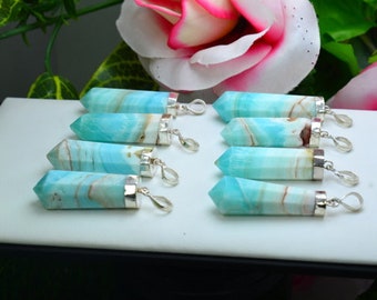May include: A collection of eight blue and white gemstone pendants with silver-colored metal caps and loops. The pendants are arranged on a white surface, with a blurred background of flowers and greenery.