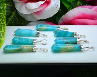 May include: Eight blue-green gemstone pendants with silver-colored metal accents are displayed on a white surface. The pendants have a rectangular shape with a pointed end. The background features pink and white flowers and green foliage.