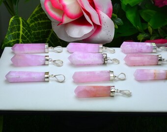 May include: A collection of pink crystal pendants with silver-colored metal caps and loops. The crystals vary in shades of pink, from light to deeper hues. The pendants are displayed on a white surface, with a floral background.