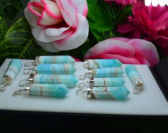 May include: A collection of blue and white gemstone pendants with silver-colored findings. The pendants are hexagonal in shape and arranged on a white surface. Pink and white flowers are in the background.