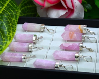 May include: A collection of pink quartz crystal pendants with silver-colored metal caps and loops. The crystals are displayed in a black tray with white compartments, with a green leaf and pink flower in the background.