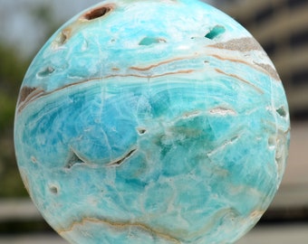May include: A polished, spherical gemstone with a striking blue and turquoise color palette. The orb features natural patterns and inclusions, with bands of brown and white. The gemstone is mounted on a light yellow base, showcasing its unique texture and color variations.