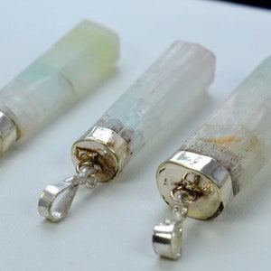 Può includere: Tre ciondoli con pietre preziose con cappucci e anelli in metallo color argento. Le pietre preziose sono rettangolari, con un aspetto traslucido e sfumature di verde, bianco e marrone. I ciondoli sono esposti su una superficie bianca.