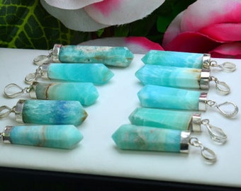 May include: A collection of turquoise-colored gemstone pendants with silver-colored metal findings. The pendants are cylindrical with pointed ends and display a range of blue and green hues. The pendants are arranged on a white surface.