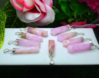 May include: A collection of pink gemstone pendants with silver-colored metal loops. The pendants are cylindrical with variations in color, from light pink to a deeper rose hue. They are arranged on a white surface, with flowers in the background.
