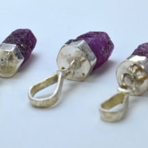 May include: Five raw ruby pendants. Each pendant features a deep purple ruby stone set with a silver-coloured metal cap and loop. The pendants are arranged in a row against a white background.