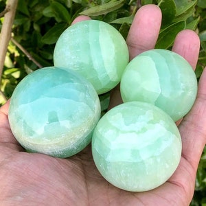 May include: Four polished, spherical stones in shades of green and turquoise are held in an open hand. The stones have a smooth, glossy surface and vary slightly in colour, with some showing subtle banding. The background is blurred green foliage.