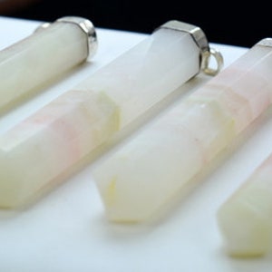 Könnte beinhalten: Vier längliche, facettierte Kristallanhänger mit silberfarbenen Kappen. Die Kristalle sind überwiegend weiß mit dezenten rosa und gelben Adern. Die Anhänger sind in einer Reihe auf einer weißen Oberfläche angeordnet.
