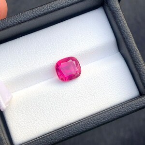 Puede incluir: Un rubí de talla cojín, exhibido en una caja de joyería negra con interior blanco. El rubí muestra un color rosa intenso y es el punto focal de la imagen. La caja es sostenida por una persona.