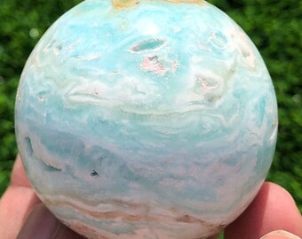 May include: A spherical gemstone in shades of light blue, white, and tan. The polished surface displays a swirling pattern, with variations in color and texture. The sphere is held in a hand against a blurred green background.