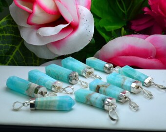 May include: A collection of turquoise-colored gemstone pendants with silver-colored metal caps and loops. The pendants are cylindrical and have a polished, smooth surface. The background features pink and white flowers.