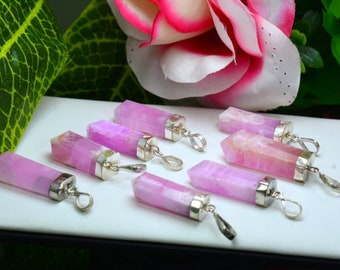 May include: A collection of pink gemstone pendants with silver-colored metal caps and loops. The pendants are rectangular with faceted tops, and are arranged on a white surface. A pink and white flower and green leaves are in the background.